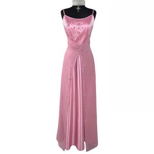 Könnte beinhalten: Ein hellrosa Abendkleid mit Spaghettiträgern, präsentiert auf einer schwarzen Schaufensterpuppe. Das Oberteil ist mit glitzernden Details verziert. Der Rock hat einen Schlitz vorne und zeigt ein Satinfutter. Eine weiße Choker-Halskette mit Kreuzanhänger ist ebenfalls zu sehen.