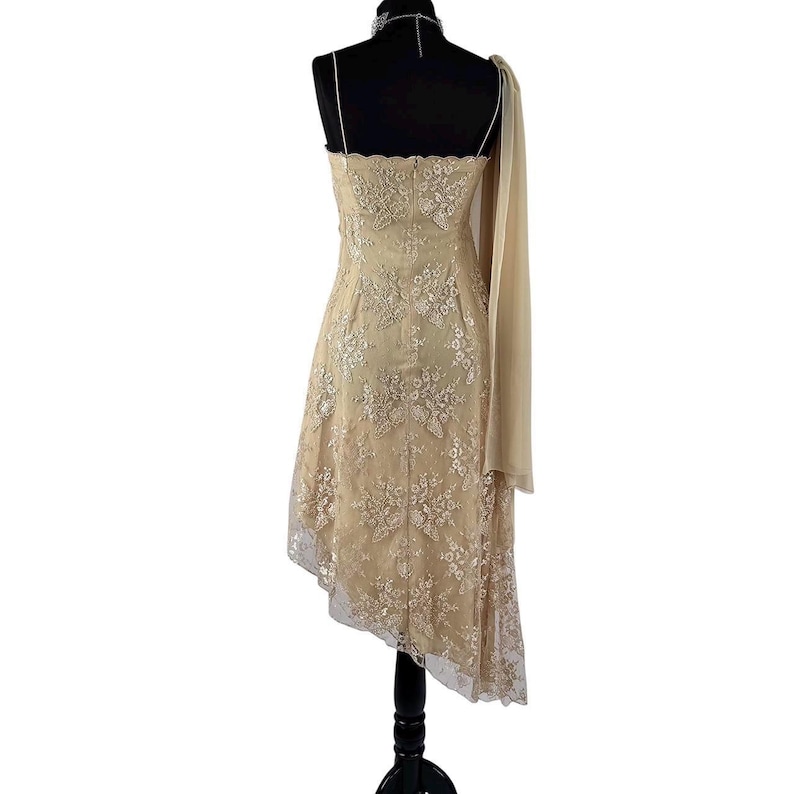 Puede incluir: Un vestido de fiesta beige asim&eacute;trico con una superposici&oacute;n de encaje floral. El vestido presenta tirantes finos y un detalle de bufanda transparente y fluida. El vestido se exhibe en un maniqu&iacute; negro.