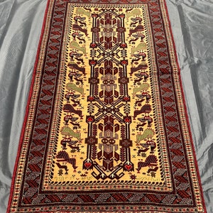 Może przedstawiać: Prostokątny beżowy dywan ze szczegółowym wzorem. Dywan ma centralny wzór z geometrycznymi kształtami i stylizowanymi motywami ptaków w odcieniach brązu, czerwieni i zieleni. Obramowanie z powtarzającymi się wzorami otacza centralny wzór. Dywan jest prawdopodobnie wykonany z wełny.