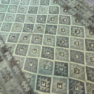 Könnte beinhalten: Ein rechteckiger Teppich mit geometrischem Muster in Grün-, Grau- und Brauntönen. Das Design zeigt sich wiederholende Quadrate und Rauten. Der Rand hat ein detailliertes, strukturiertes Muster.