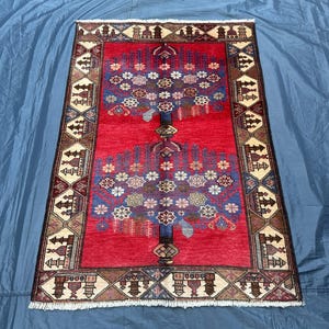 May include: A red rug with a traditional Persian design featuring two stylized trees with floral motifs. The rug has a cream and brown border with geometric patterns.