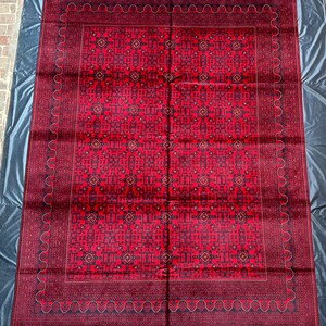 Op de afbeelding: Een rood tapijt met een geometrisch patroon in zwart en wit. Het tapijt heeft een franje rand.