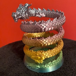 Peut inclure: Une pile de quatre figurines de dragon argentées, roses, dorées et vertes. Chaque dragon est enroulé en cercle, la tête du dragon du haut étant tournée vers le spectateur.