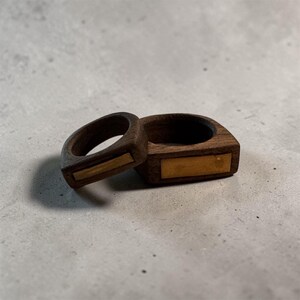 May include: Two dark brown wooden rings featuring a rectangular gold-toned metal inlay on the front surface. The rings have a smooth, polished finish and a thick, rounded band design, showcasing the natural grain of the dark wood material.