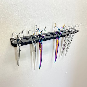 May include: A black acrylic shear holder mounted on a white wall, displaying several pairs of professional hair shears. The shears are silver and some have iridescent rainbow handles. The holder has multiple slots to hold the shears.