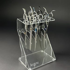 May include: A clear acrylic display stand holding several pairs of professional hair shears. The shears are silver and black, with various handle designs. The stand has the brand name "XIZZORZ" printed on the base.