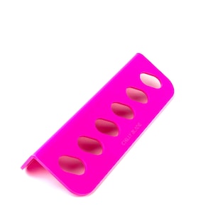 May include: A bright pink, angled kitchen tool with five oval cutouts. The text "CHILLY BLADE" is printed on the side. The tool is designed to sit on a surface and has a folded edge.