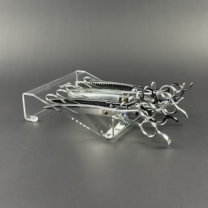 May include: A clear acrylic organizer holding several pairs of professional hair shears. The shears are silver and black, with various blade and handle designs. The organizer is angled and has a minimalist design.