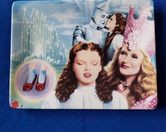 May include: A ceramic plate featuring a colorful image from the Wizard of Oz movie. The plate shows Dorothy, the Tin Man, and the Wicked Witch of the West. The plate also features Dorothy's ruby red slippers.