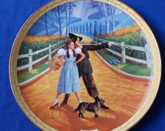 May include: A ceramic plate featuring a colorful illustration of Dorothy and the Scarecrow from the Wizard of Oz walking down the yellow brick road. The plate has a gold rim and the words "The Wizard of Oz" in a gold banner.