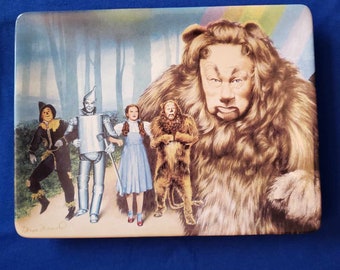 May include: A ceramic plate featuring a colorful illustration of the characters from the Wizard of Oz. The Cowardly Lion, Dorothy, the Tin Man, and the Scarecrow are walking down a yellow brick road. The Cowardly Lion is in the foreground, with Dorothy, the Tin Man, and the Scarecrow behind him. The background is a forest with a rainbow in the sky.