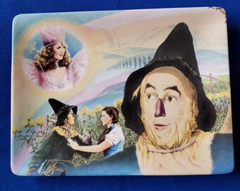 May include: A ceramic plate with a colorful image of the Scarecrow from the Wizard of Oz. The Scarecrow is wearing a straw hat and a brown shirt. He is standing in a field of yellow flowers. Dorothy is standing next to him, and the Wicked Witch of the West is in the background.