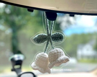 May include: A pair of white crocheted bells with green crocheted leaves hanging from a rearview mirror in a car. The text "@mellowstitchco" is visible in the bottom right corner of the image.