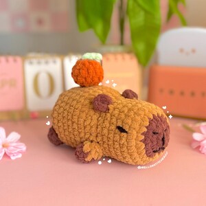 May include: A handmade, crocheted capybara plushie in shades of brown and tan, with a small orange pumpkin on its back. The plushie has a dark brown face and small, dark brown ears and feet. The text "mellowstitchesCo" is visible.