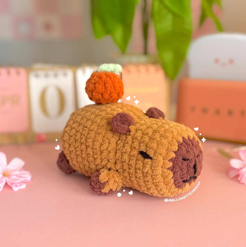 May include: A handmade, crocheted capybara plushie in shades of brown and tan, with a small orange pumpkin on its back. The plushie has a dark brown face and small, dark brown ears and feet. The text "mellowstitchesCo" is visible.