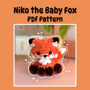 Pode incluir: Um brinquedo de raposa de crochê em tons de laranja, branco e preto, com o texto "Niko the Baby Fox PDF Pattern" acima. A raposa está sentada em uma superfície rosa. O brinquedo tem uma textura macia e fofa e uma aparência amigável.