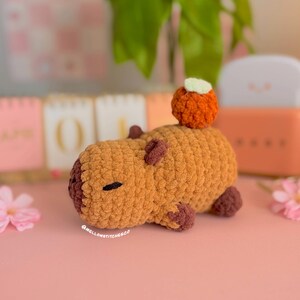 May include: A handmade crochet capybara in shades of brown and tan, with a small orange pumpkin on its back. The capybara has small, dark brown details for its eyes, ears, and feet. The text "@mellowstitches.co" is visible on the bottom left.