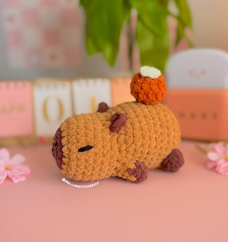 May include: A handmade crochet capybara in shades of brown and tan, with a small orange pumpkin on its back. The capybara has small, dark brown details for its eyes, ears, and feet. The text "@mellowstitches.co" is visible on the bottom left.