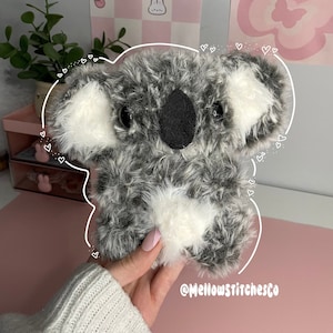 Może przedstawiać: Pluszowy miś koala z szarym i białym futrem, czarnymi oczami i czarnym nosem. Koala ma białe futro na uszach i klatce piersiowej. Widoczny jest tekst "@Mellowstitchesco".