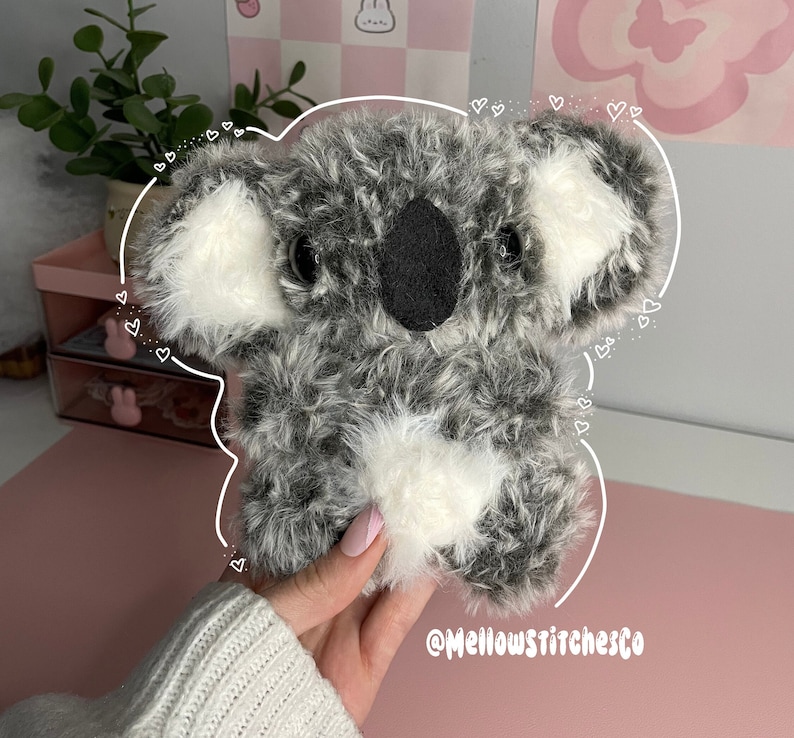 Może przedstawiać: Pluszowy miś koala z szarym i białym futrem, czarnymi oczami i czarnym nosem. Koala ma białe futro na uszach i klatce piersiowej. Widoczny jest tekst "@Mellowstitchesco".