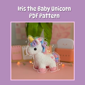 May include: A white crocheted baby unicorn with a pink, blue, and yellow mane and tail. The unicorn is standing on a pink surface with a blurry background.