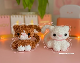 May include: Two crocheted cat plushies, one brown and white and one white with a pink ribbon. The plushies are sitting on a pink surface. @mellowstitchiesco