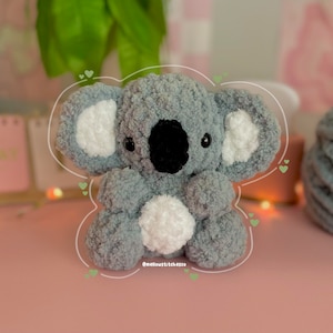 Może przedstawiać: Ręcznie robiony szary i biały pluszowy miś koala z czarnym nosem i oczami. Koala ma puszyste, teksturowane futro i biały pompon na brzuchu. Koala siedzi na r&oacute;żowej powierzchni. Widoczny jest tekst "@mellowstitchesco".