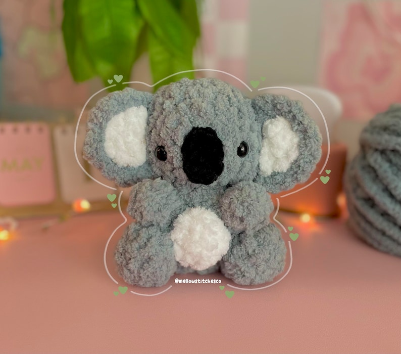 Może przedstawiać: Ręcznie robiony szary i biały pluszowy miś koala z czarnym nosem i oczami. Koala ma puszyste, teksturowane futro i biały pompon na brzuchu. Koala siedzi na r&oacute;żowej powierzchni. Widoczny jest tekst "@mellowstitchesco".