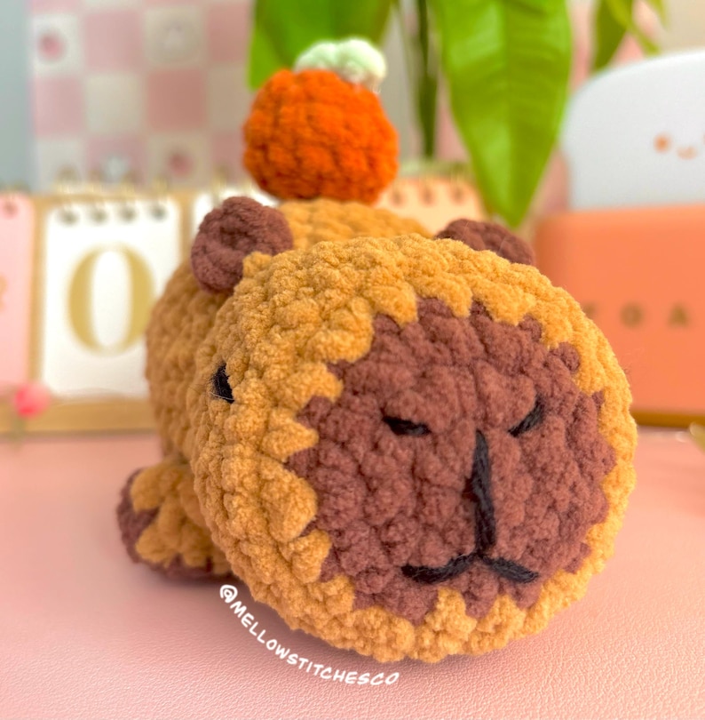 May include: A handmade crochet lion with a pumpkin on its head. The lion is primarily brown and tan, with a dark brown face and black embroidered details. The pumpkin is orange with a white stem. The text "@mellowstitchesCo" is visible.