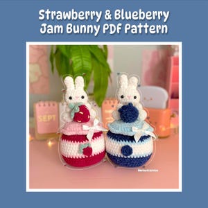 Puede incluir: Dos peluches de conejo amigurumi tejidos a crochet, uno con una fresa y otro con un arándano, sentados sobre una superficie rosa. Los conejos llevan tarros de mermelada como sombreros, el conejo de fresa lleva un tarro rosa y blanco y el conejo de arándano lleva un tarro azul y blanco. El texto "Strawberry & Blueberry Jam Bunny PDF Pattern" está en la parte superior de la imagen.