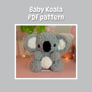 May include: A grey crocheted baby koala plush toy with white ears and a white belly. The koala is sitting on a pink surface with green hearts around it.