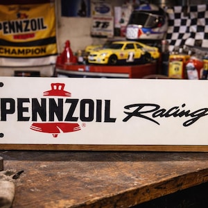 May include: A rectangular white metal sign featuring the black text PENNZOIL and the word Racing in a black script font. A red bell-shaped logo is positioned between the words. The sign has four black mounting holes in the corners.