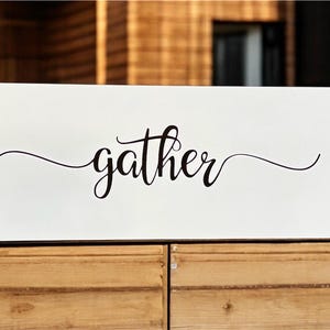 May include: A rectangular white sign with rounded corners and the word "gather" in elegant black script. The sign has four small holes, one in each corner. The sign is placed on a wooden surface.