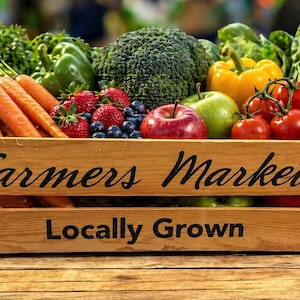 May include: A wooden crate filled with fresh produce, including carrots, strawberries, blueberries, apples, tomatoes, and radishes. The crate is labeled "Farmers Market Locally Grown."