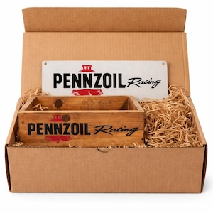 May include: A rectangular brown cardboard shipping box containing a wooden crate and a metal sign, both featuring the black and red Pennzoil Racing logo. The items are nestled in light brown wood shavings inside the open box.