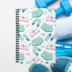 May include: A white spiral notebook with a colorful pattern of pink sports bras, teal shorts, and blue running shoes. The notebook is lying on a white surface with blue dumbbells and a blue water bottle.