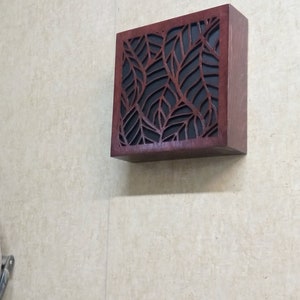 May include: A dark brown wooden wall mounted box with a leaf pattern cut out design. The box has a black background.