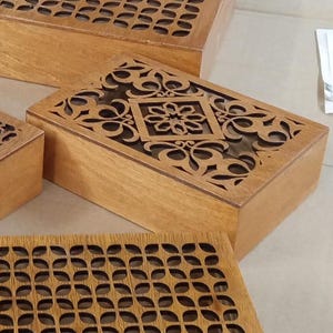 May include: Wooden boxes with intricate laser-cut designs on the lids. The boxes are rectangular and have a natural wood finish. The lids feature floral and geometric patterns, adding a decorative element to the storage containers.