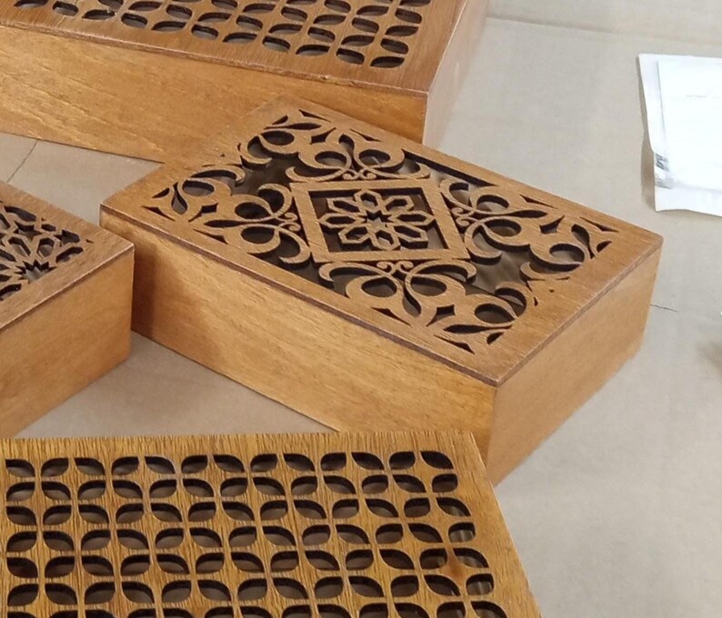 May include: Wooden boxes with intricate laser-cut designs on the lids. The boxes are rectangular and have a natural wood finish. The lids feature floral and geometric patterns, adding a decorative element to the storage containers.