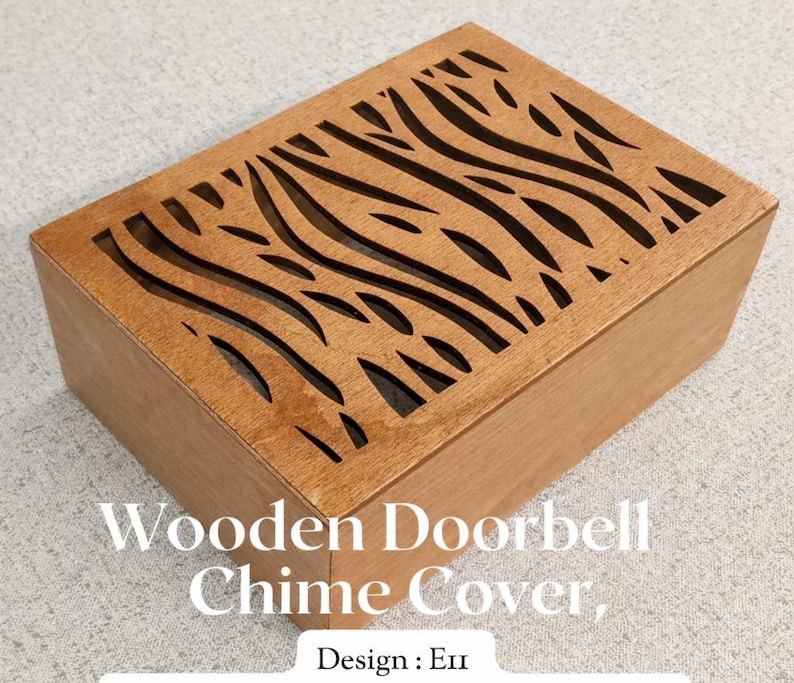 May include: A rectangular wooden doorbell chime cover with a laser-cut design on the top. The cover is made of light brown wood and features a series of curved, wave-like cutouts. The text "Wooden Doorbell Chime Cover" and "Design: En" are visible.
