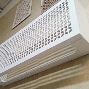 May include: A white, rectangular air vent cover with a decorative, leaf-shaped pattern. The cover has a handle-like cutout on the bottom and is mounted on a beige wall. Additional decorative panels are visible.