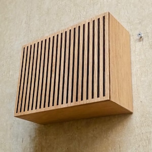 May include: A rectangular wooden box with vertical slats on the front, mounted on a light-coloured wall. The box is made of light-coloured wood, and the slats create a striped pattern. The box is mounted with a screw.