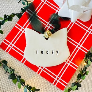 May include: White ceramic Christmas ornament shaped like a cat with the name "Rocky" written on it. The ornament is on a red and white plaid background with a green ribbon and a gold bell.