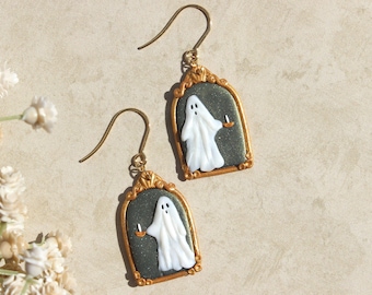 May include: A pair of gold-toned earrings with a black and green background featuring a white ghost holding a candle. The ghost is framed in a gold-toned arch.