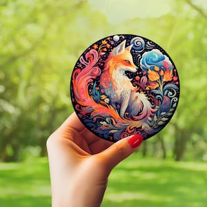 Puede incluir: Un posavasos redondo y colorido con un zorro estilizado con un cuerpo blanco y naranja, rodeado de patrones florales en espiral en tonos azules, rojos y amarillos.