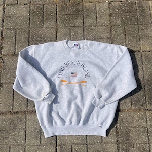 May include: A light gray sweatshirt with the text "Long Beach Island" in a circular design. The design includes an American flag and crossed oars. The sweatshirt is made of a soft material and has a crew neck.