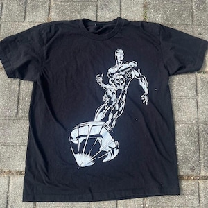 May include: Black t-shirt featuring a white graphic of a muscular figure emerging from a stylized, spherical object. The design is detailed with shading, creating a dynamic and artistic look. The shirt is made of a soft material.