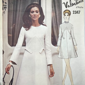 Könnte beinhalten: Ein Vintage-Schnittmuster von Vogue Grand Couturier mit einem weißen Kleid-Design von Valentino d'Italie. Die Schnittmuster-Nummer lautet 2347. Das Schnittmuster zeigt eine Frau in einem weißen Kleid mit V-Ausschnitt und einem taillierten Oberteil. Das Kleid hat einen ausgestellten Rock und lange Ärmel. Das Model hält eine schwarze Handtasche.