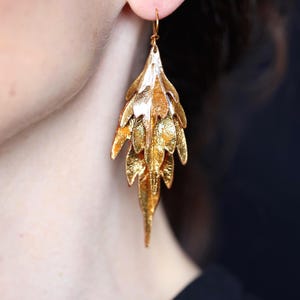 May include: A gold-toned drop earring featuring a layered, multi-leaf design with a textured, metallic finish. The earring hangs from a simple hook wire and consists of several overlapping, pointed leaf-shaped elements in a cascading arrangement.