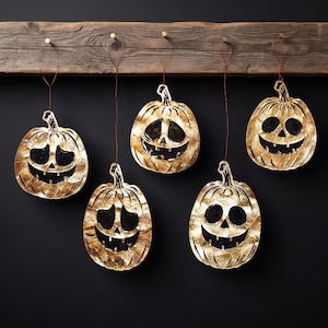 May include: Five hanging pumpkin ornaments with a metallic gold finish and black cut-out facial features. Each pumpkin is suspended by a thin red string from a horizontal wooden plank. The pumpkins display various carved expressions.